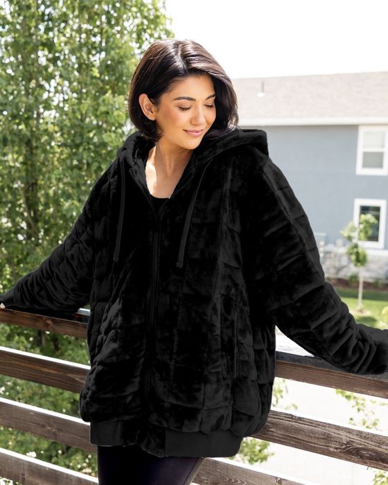 A person stands outdoors on a balcony, wearing an oversized plush fur jacket with a hood