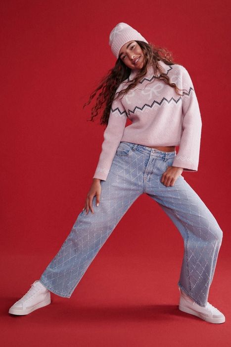 A smiling person dressed in a patterned sweater, beanie, and wide-leg jeans, striking a playful pose against a simple background