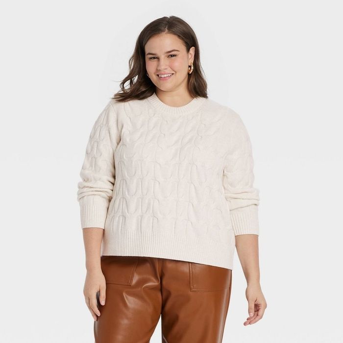 A person smiles, wearing a cable knit sweater and brown leather pants, facing the camera.