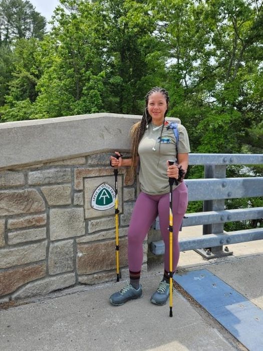 A reviewer with trekking poles stands by a trail sign, wearing athletic leggings, a top, and a backpack.
