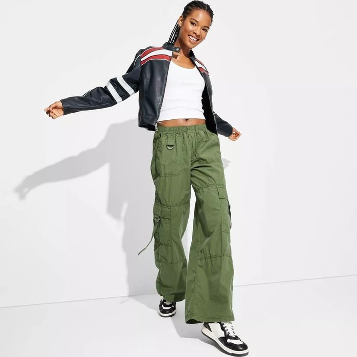 A smiling model wears a white tank top, a black and red-striped jacket, green cargo pants, and sneakers