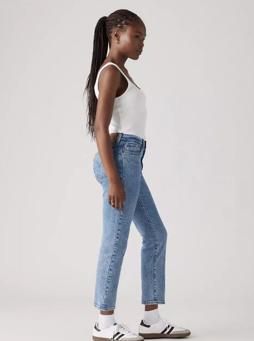 A person in a white tank top, blue jeans, and sneakers, striking a profile pose with a cool, casual vibe.