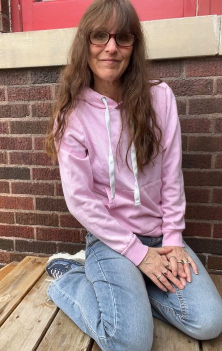 A reviewer seated on wooden steps, wearing a hoodie and jeans, smiling with long hair and glasses, set against a brick wall backdrop.