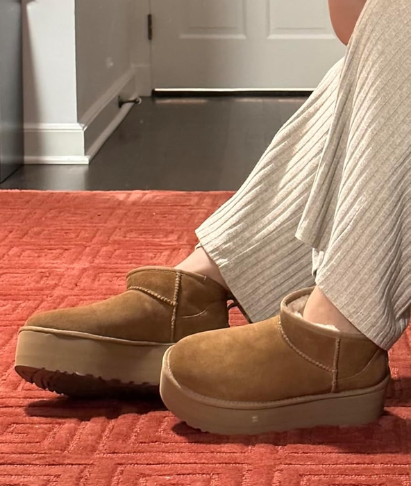 A person taking a mirror selfie in beige pleated pants and tan platform slippers on an orange textured rug
