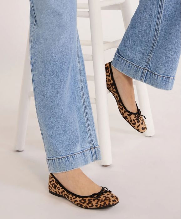 A person wearing blue jeans and leopard-print ballet flats, standing near a white stool. Featured in a shopping article