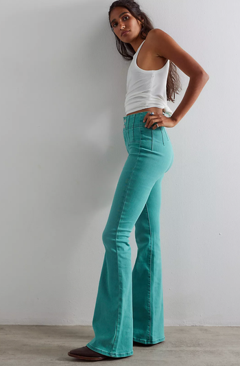 A person wearing flared jeans and a sleeveless top strikes a pose against a simple wall, displaying casual style.