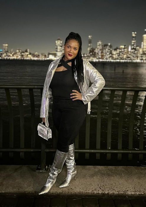 A person confidently stands by the riverside at night with a city skyline in the background, wearing a silver jacket, matching knee-high boots, a black top, and pants while holding a purse
