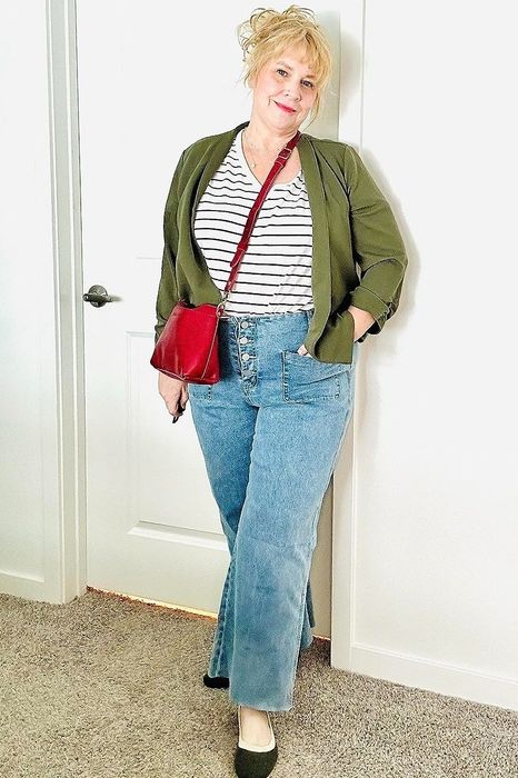 A reviewer dressed in a striped t-shirt, green blazer, high-waisted blue jeans, black shoes, and a red crossbody bag, posing near a door in a well-lit room.