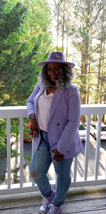 A person standing on a balcony, showcasing a chic lavender blazer, a matching hat, a white top, distressed jeans, and sneakers, set against a backdrop of lush trees.