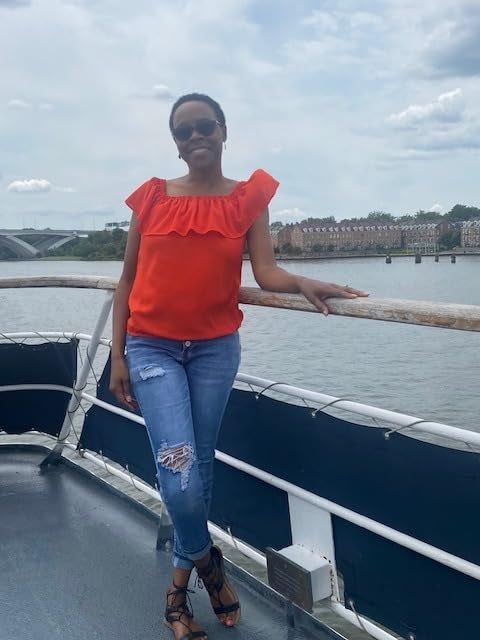 Someone relaxing on a boat, sporting a red off-shoulder top, ripped jeans, and sunglasses, with a serene river and buildings in the distance.
