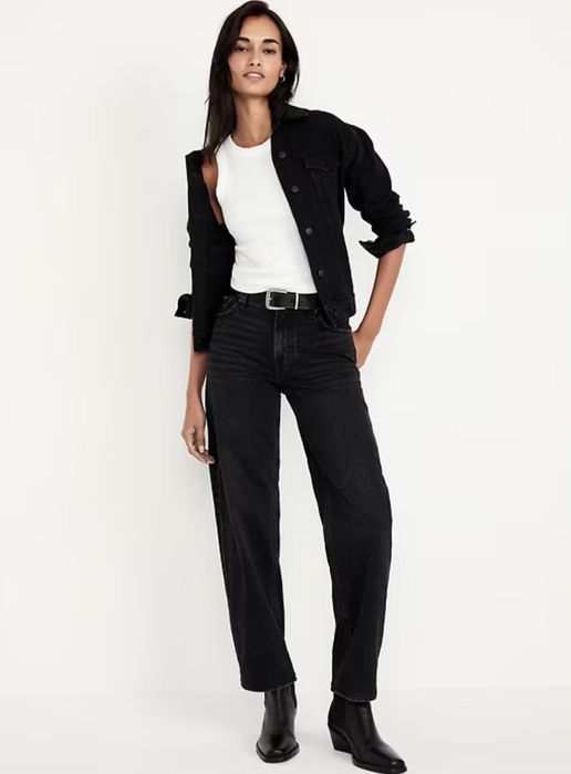A person confidently poses in a black denim jacket, white top, black jeans, and black boots, showcasing a chic, all-black ensemble.