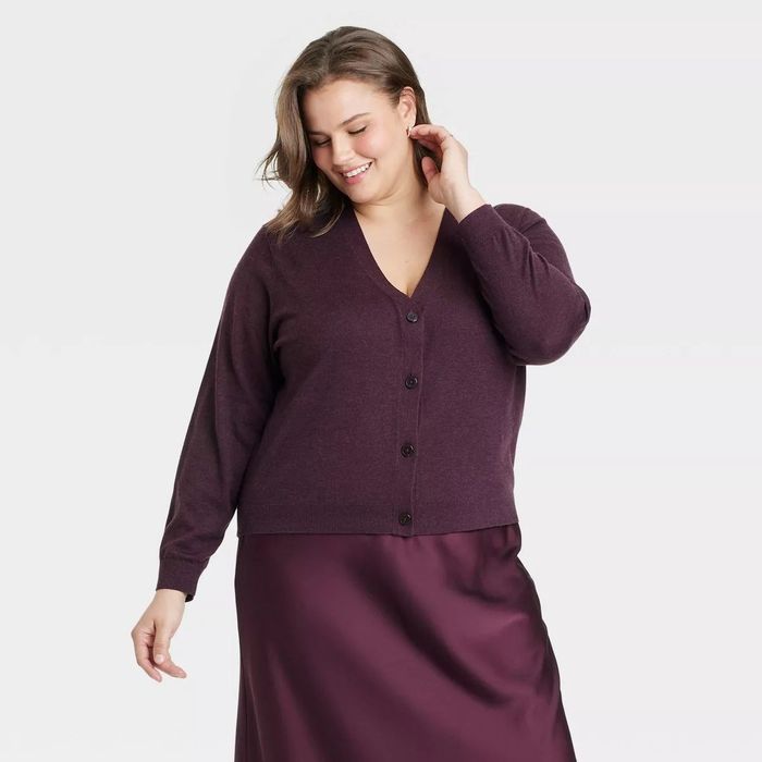 A model wears a long-sleeve cardigan paired with a satin skirt, smiling and looking downward.
