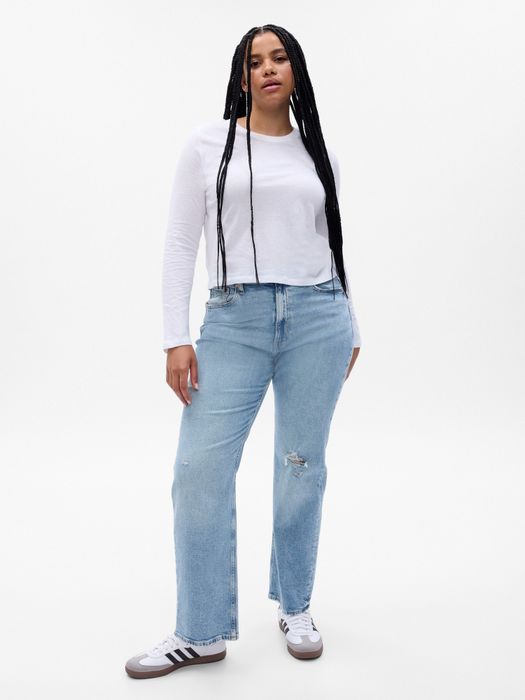 A model with long braids, dressed in a white long-sleeve top, light blue jeans, and sneakers.