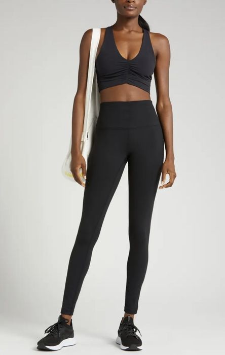 A model is dressed in black high-waisted leggings, paired with a black sports bra, black sneakers, and is holding a white tote bag.