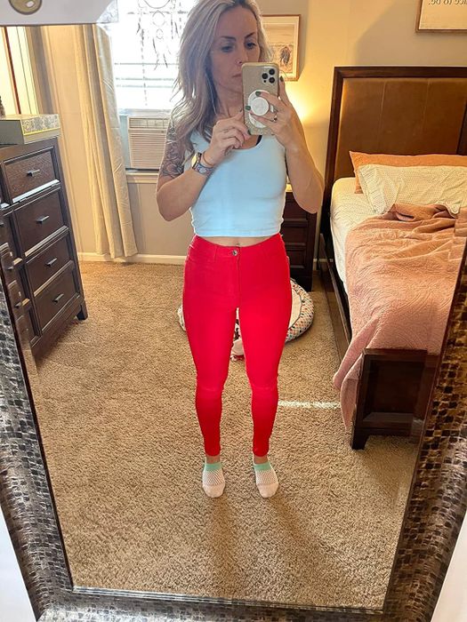 A reviewer takes a mirror selfie, showcasing a white tank top paired with vibrant red jeans.