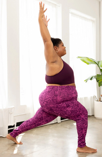 A person practicing yoga in a well-lit room, wearing a chic workout set with a one-shoulder top and patterned leggings.