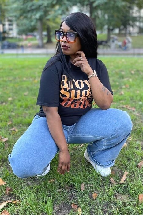 A reviewer crouches on the grass in a park, wearing a graphic tee, jeans, and large glasses, gazing to the side.
