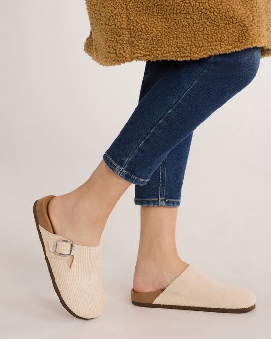 A person wearing snug slip-on clogs paired with cropped jeans, standing with a fuzzy coat in view