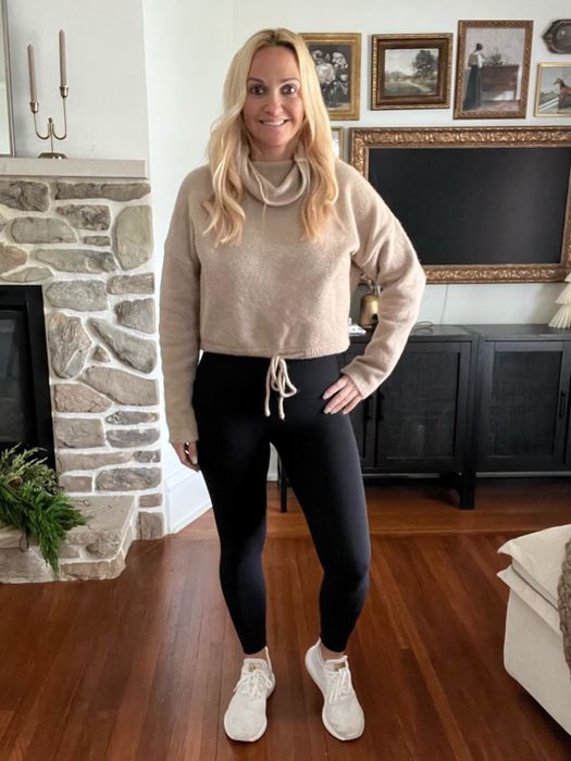 A reviewer dressed in a casual outfit featuring a beige sweater, black leggings, and white sneakers, standing in a living room with a stone fireplace and framed pictures on the wall.