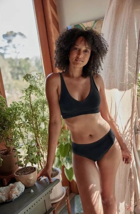 A model in a relaxed black bralette and matching underwear set poses beside indoor plants