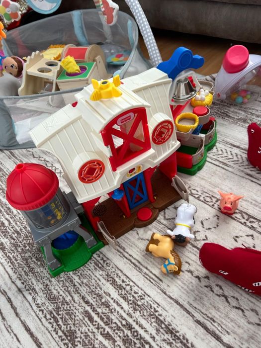 A children's farm playset featuring animals and farmer figures, set on a carpet. The toy barn and silo are at the center, surrounded by miniature figures and various other toys.
