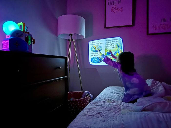 A child in pajamas enjoys a bedtime story projected on the wall, with the room softly lit by a comforting nightlight.