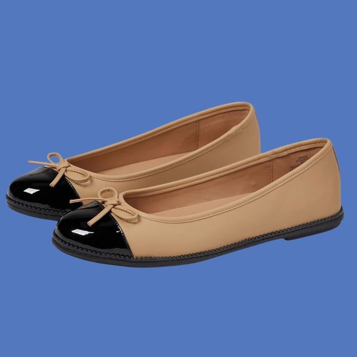 Beige ballet flats with black glossy toe caps and a small bow detail, set against a blue background.