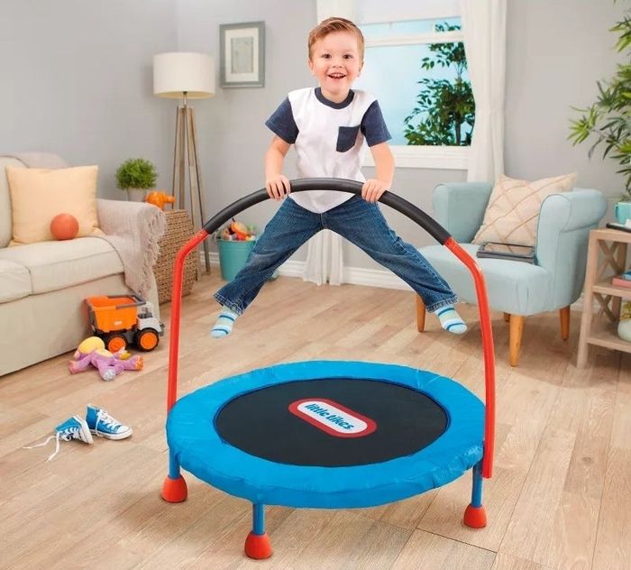 A child happily bounces on a compact indoor trampoline with handlebars, surrounded by toys in a cozy living room