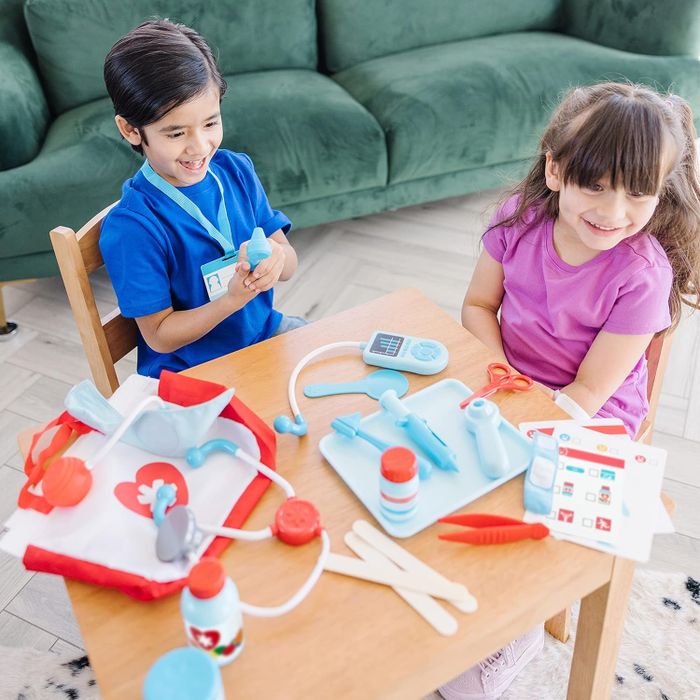 Two children joyfully play with a toy medical kit on a wooden table, complete with a stethoscope, syringe, and various doctor tools, sparking imaginative play.