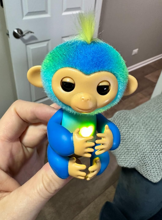 A hand holds a small, fuzzy toy monkey with large eyes and a glowing light in its chest, showcasing its interactive feature.