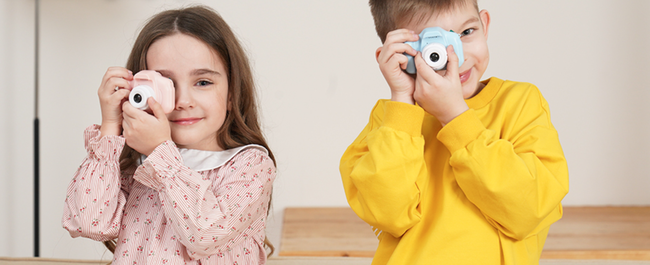 Two children, a girl wearing a patterned dress and a boy in a long-sleeve shirt, are each holding a toy camera.