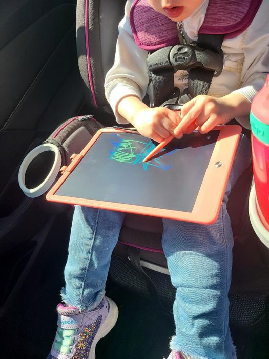 A child drawing on the tablet while seated in a car, holding the stylus with a pair of jeans and sneakers visible in the scene.