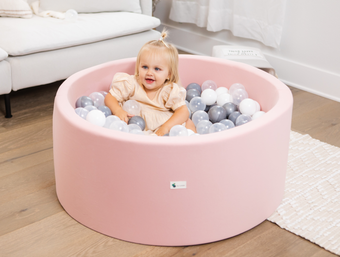 A toddler sits in a round ball pit filled with black, white, and gray balls, grinning. The background looks cozy and inviting, resembling a comfortable living room.