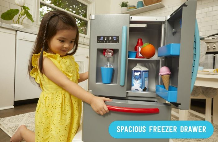A child plays with a toy kitchen set's fridge, which features a spacious freezer drawer filled with plastic food items like milk and ice cream.