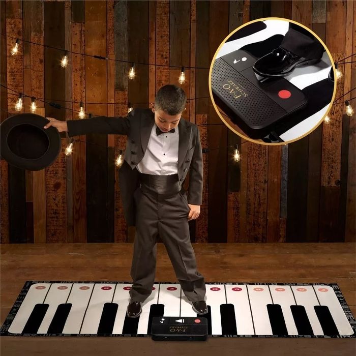 A child dressed in a tuxedo and holding a top hat plays on a large piano keyboard mat, featuring a control unit with various settings