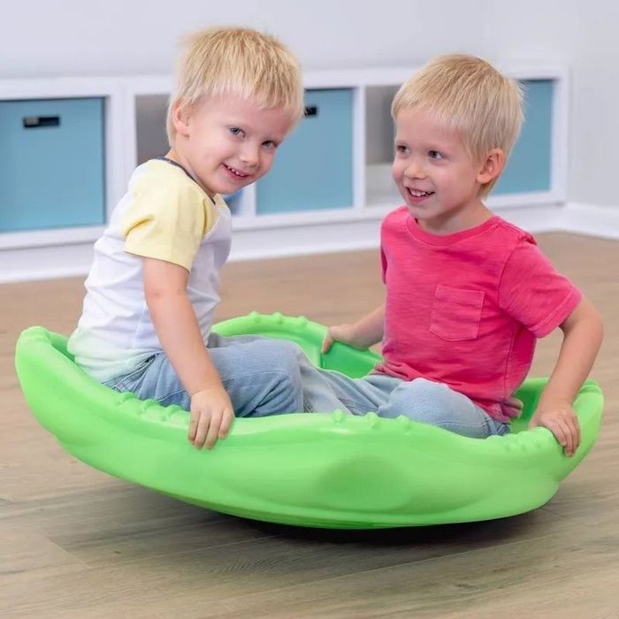 Two young kids sit together on a green spinning toy indoors, smiling and enjoying their playtime