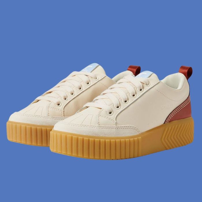 A pair of beige Sorel sneakers featuring thick gum soles, white laces, and red accents on the heels, set against a blue background.