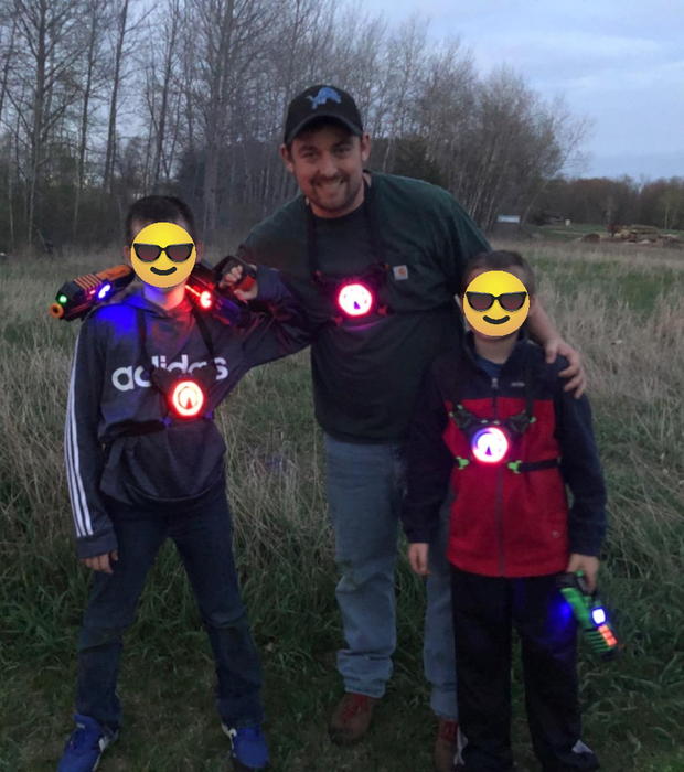 Three people grinning outdoors, geared up in laser tag equipment with glowing targets. Two kids and an adult man in casual clothes, each holding a laser gun