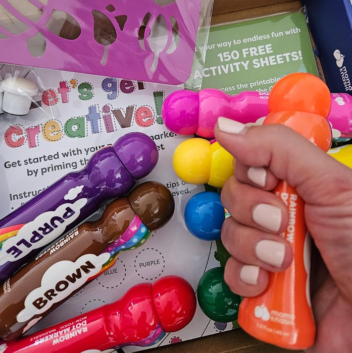 A colorful activity kit featuring paint markers, an instruction booklet, and a warning about the dangers of plastic packaging. A hand is holding an orange marker.