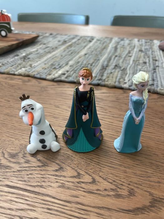 Three Frozen character figurines: Olaf the snowman, Anna in a green gown, and Elsa in a sparkling ice-themed dress, placed on a wooden table.