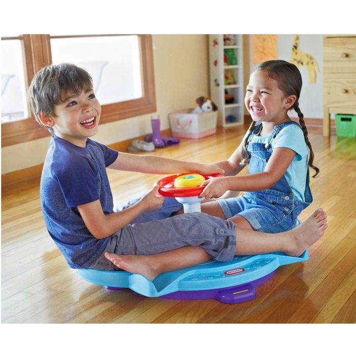 Two kids smile as they sit on a toy spinner in a playroom. The girl is in overalls, and the boy wears a t-shirt and shorts