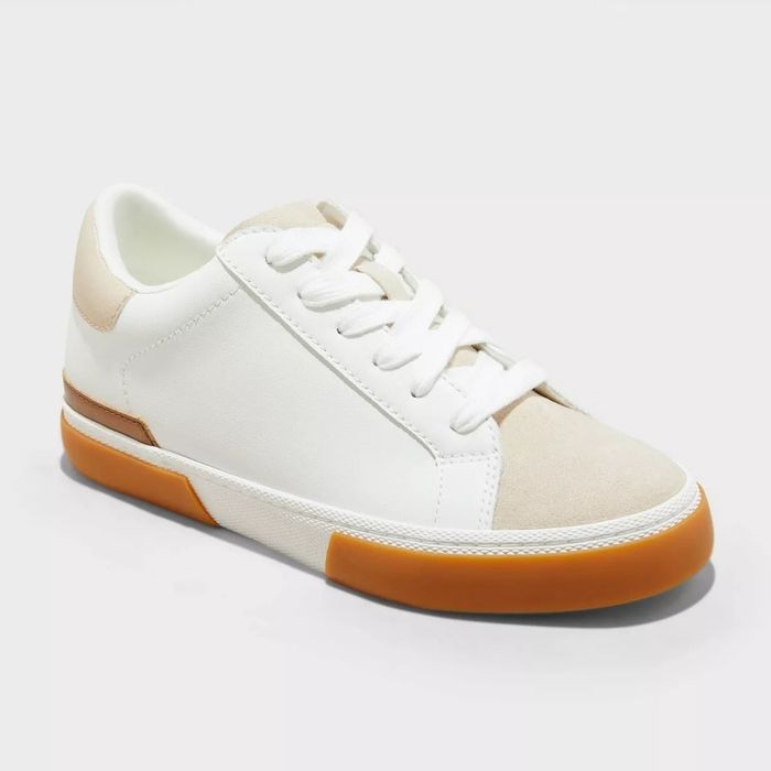 A stylish sneaker with a white body, gum sole, and light beige suede accents is shown. The laces are white, and the overall design is sleek and modern.