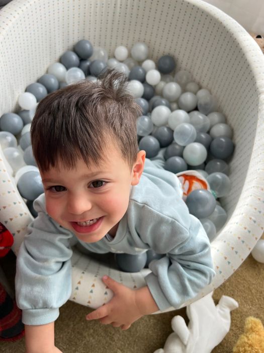 A child smiles happily in a ball pit filled with plastic balls, including shades of gray and white. The environment feels playful and laid-back.