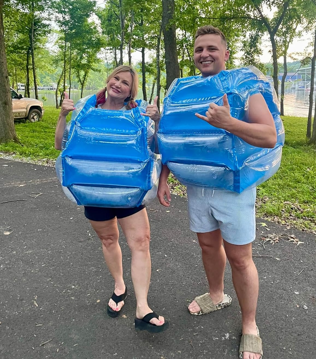 Two people outdoors wearing inflatable bumper vests, laughing and posing for the camera. A backdrop of trees and their casual summer outfits create the perfect setting.