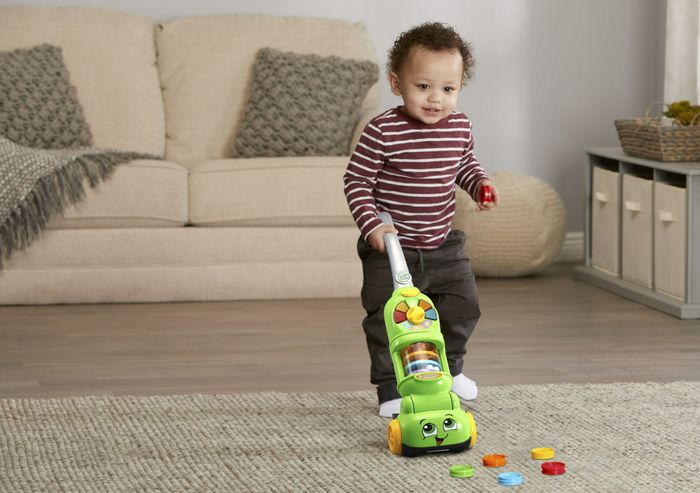 A boy happily pushes a vacuum, mostly green, as it picks up small toy pieces scattered around.
