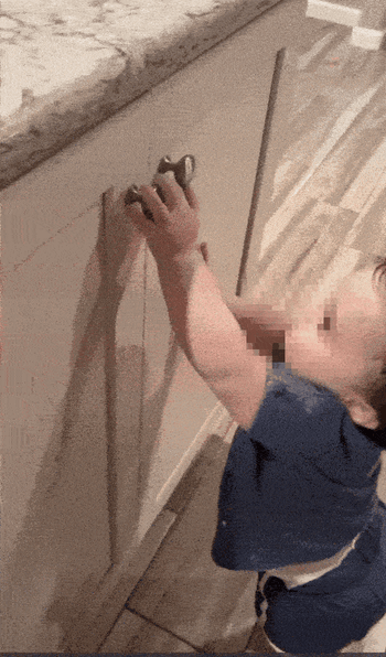 a toddler in a blue shirt attempting to open a kitchen cabinet, with safety locks effectively preventing access