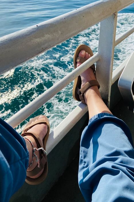 Reviewer showcasing the sandals with velcro straps in light brown, paired with blue pants, on a boat.