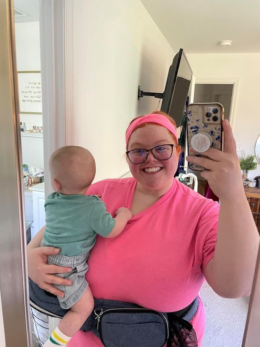 a person wearing a TushBaby smiles at their reflection in the mirror while holding their baby