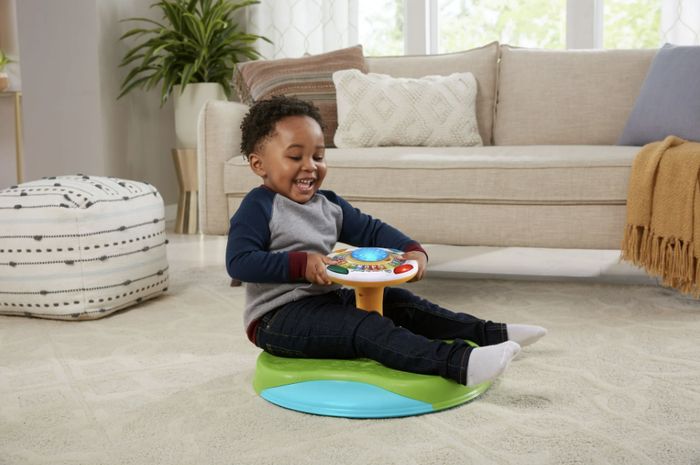 A child joyfully spins on a Sit 'n Spin toy in a living room, with a couch and pouf visible in the background.