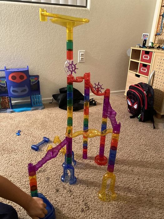A vibrant marble run toy setup rests on a carpeted floor inside a room, with a backpack and other toys visible in the background.